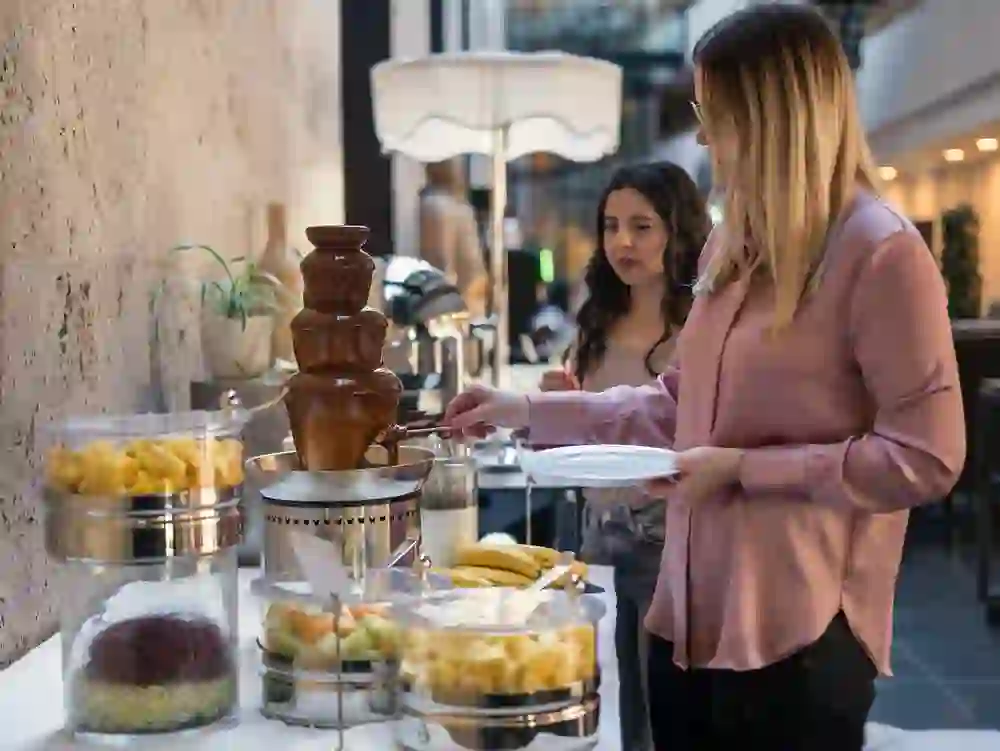 Zwei Frauen bedienen sich an einem Dessertbuffet mit Schokoladenbrunnen in einem modernen Innenraum. Die Frau im Vordergrund hält einen Teller und taucht ein Stück Frucht in den fliessenden Schokoladenbrunnen, während im Hintergrund verschiedene frische Früchte wie Ananas, Melone und Bananen bereitstehen. Die Atmosphäre ist entspannt und einladend – passend zu einem Brunch oder einem gehobenen Hotelbuffet.