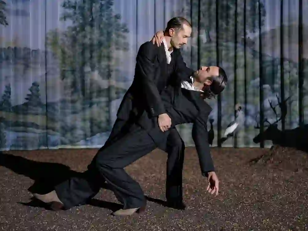 Two people in suits strike a dramatic dance pose on a dark gravelled surface in front of a forest backdrop.