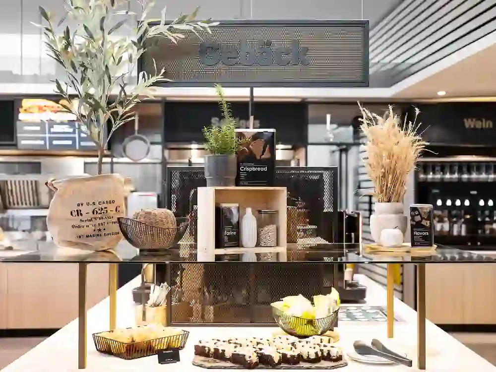 Sur cette photo, on peut voir une zone de pâtisserie d'un buffet présentée avec style. Au centre se trouve une table disposée de manière décorative avec différentes pâtisseries, du pain croustillant et des snacks sucrés. La scène est décorée avec des détails soignés :  Un grand panneau portant l'inscription "pâtisseries" est suspendu au-dessus du buffet.  Sur la table se trouvent entre autres :  Une corbeille de pain croustillant (étiqueté "Crispbread"), Une petite corbeille de crackers ou de biscuits préemballés, Un plateau avec des brownies ou des parts de gâteau, Des éléments décoratifs comme un petit olivier, une corbeille de miches de pain, des céréales sèches dans un vase et des sacs en jute rustiques.  En arrière-plan, on distingue d'autres zones comme "Vin" et "Spécial", qui font probablement partie d'un concept de buffet ou de restauration plus important.  L'ensemble donne une impression de qualité, de réflexion et de modernité, avec un mélange de décoration naturelle et de chic industriel. La présentation invite clairement à la dégustation.