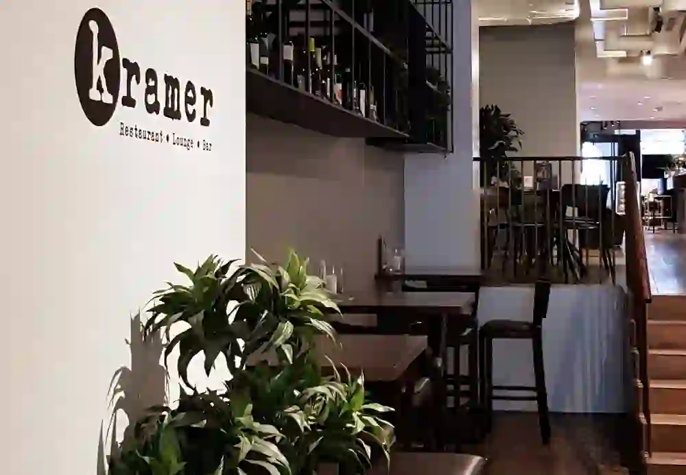 The picture shows the interior of a restaurant called Kramer, which operates as a restaurant, lounge and bar, as the wall labelling reveals. Plants adorn the foreground, while in the background a staircase leads upstairs where more seating can be seen. The style is modern and inviting, with a calm and relaxed atmosphere. The dark furniture contrasts pleasantly with the light-coloured floor and walls.