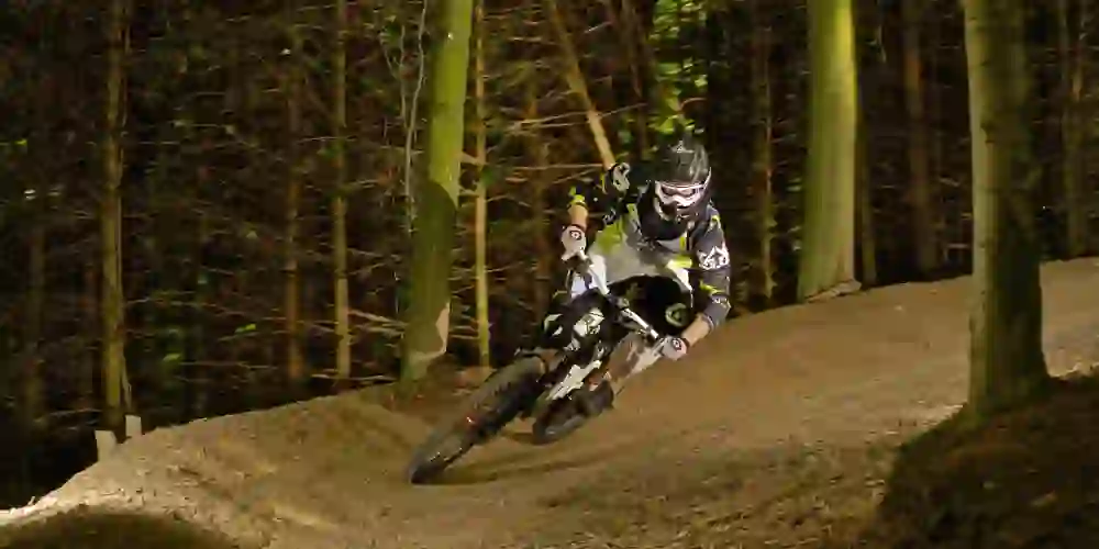 Im Fokus des Bildes ein Mountainbiker, der sich konzentriert und dynamisch eine beleuchtete Downhill-Strecke hinuntermanövriert, eingebettet in die nächtliche Ruhe eines Waldes.