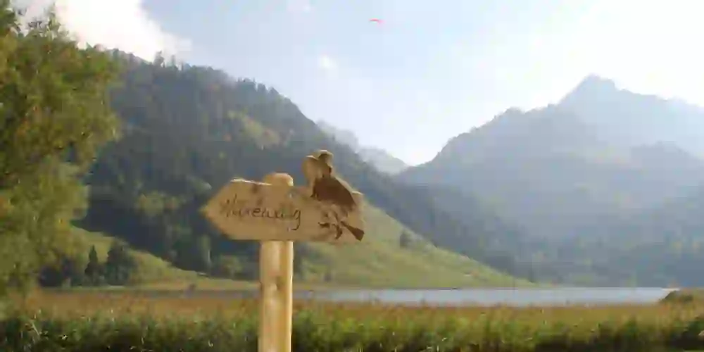 L'image montre un panneau indicateur en bois avec l'inscription "Hexewääg" et une figure de sorcière dessus. En arrière-plan, on distingue un paysage pittoresque avec un lac, des prairies vertes et des collines boisées. Dans le ciel, on aperçoit un parapentiste. La scène semble calme et contemplative et invite à une promenade détendue sur le "chemin des sorcières".