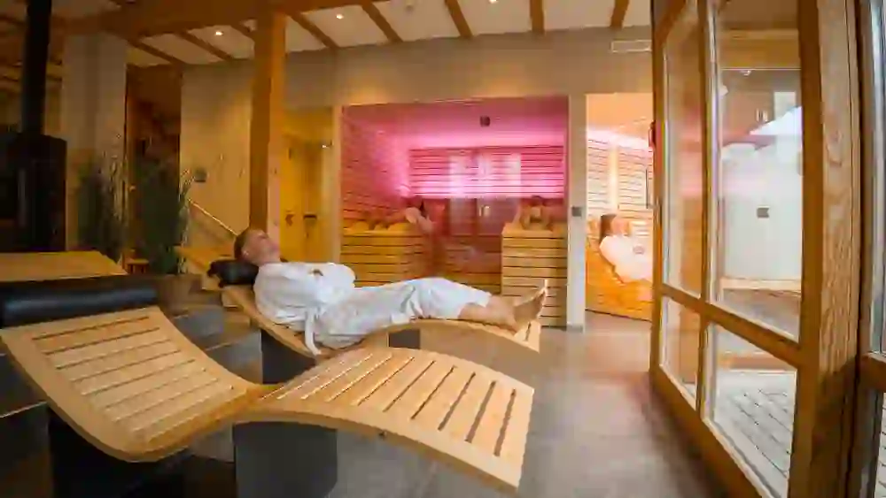 In einem warmen, holzgetäfelten Wellnessbereich entspannt sich eine Person auf einer Liege, die Augen geschlossen, die Ausstrahlung friedvoll. Durch das große Fenster scheint sanftes Tageslicht herein und bietet einen Blick in die entspannende Atmosphäre der Sauna im Hintergrund.