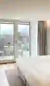 Das Bild zeigt ein helles und modern eingerichtetes Hotelzimmer. Ein großes, komfortables Bett steht im Zentrum, flankiert von stilvollen Möbeln und Pflanzen. Natürliches Licht flutet durch die Glastüren, die auf einen Balkon mit Aussicht auf eine Stadtlandschaft führen. Die Atmosphäre wirkt einladend, ruhig und komfortabel. Das Design ist schlicht und elegant, was eine entspannende Stimmung vermittelt, perfekt für Erholung und Ruhe.