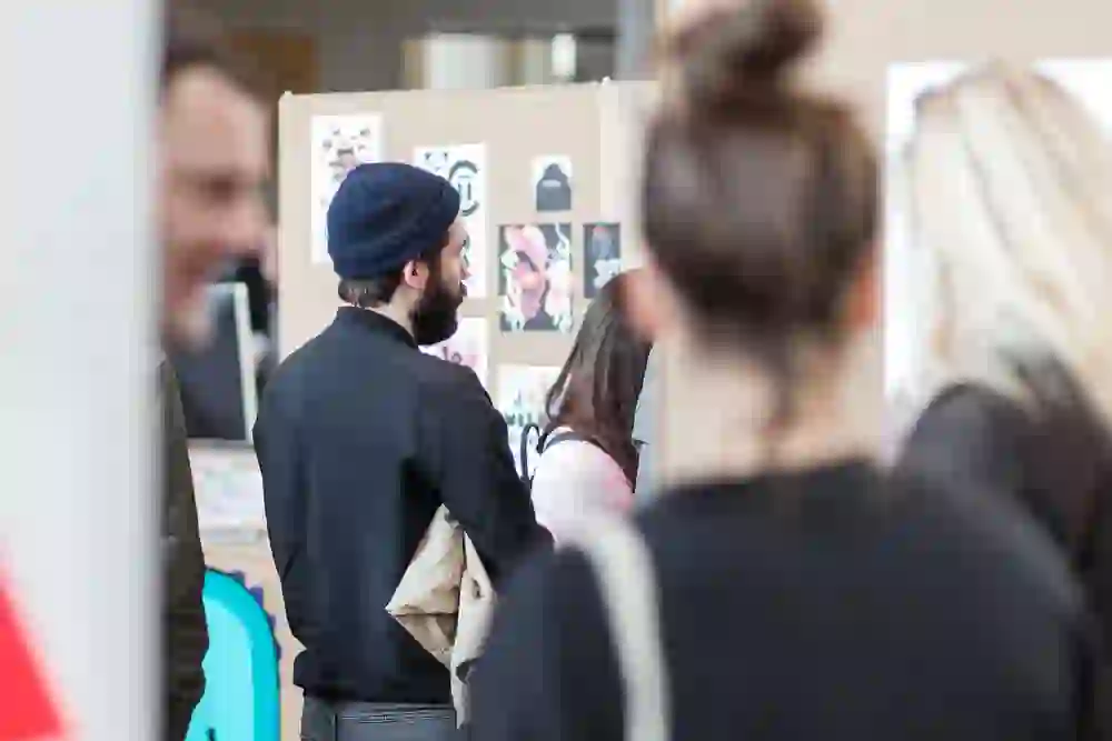 La photo montre des visiteurs qui participent à une exposition dans le cadre du Design Festival Bern. On les voit en train d'avoir des conversations animées tout en regardant les œuvres exposées. L'atmosphère est empreinte de créativité et d'échange, typique d'un festival qui célèbre le design et la communauté.