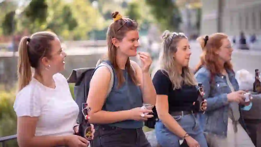 Das Foto zeigt vier Frauen, die lächelnd und unterhaltend an einem Geländer stehen. Sie halten Bierflaschen und scheinen eine angenehme Zeit zu geniessen. Ihre entspannte Körperhaltung und die sichtbare Interaktion deuten auf eine freundschaftliche und ungezwungene Atmosphäre hin. Im Hintergrund ist eine städtische Flusskulisse zu erkennen, die zum gemütlichen Ambiente beiträgt.