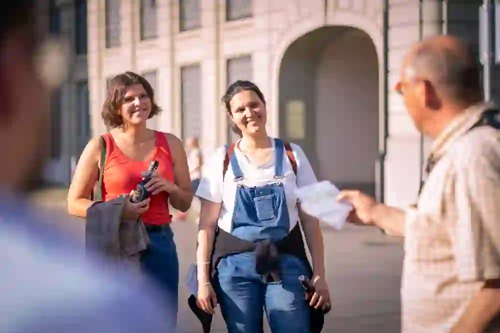 La photo montre deux femmes qui suivent l'édition Fübi du Berner Biertour ; l'une tient une bouteille de bière et un programme, tandis que l'autre sourit et regarde directement l'appareil photo. Elles sont habillées en été et semblent détendues et intéressées. Une autre personne, le guide de la ville, est visible au premier plan, dos à la caméra.