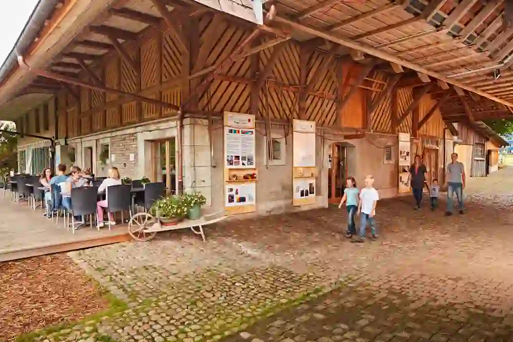 Un'atmosfera rilassata circonda i visitatori della Spycher-Handwerk AG che siedono comodamente nell'area esterna coperta. Le famiglie passeggiano ammirando l'architettura rustica e la mostra informativa inserita nell'ambiente rurale.