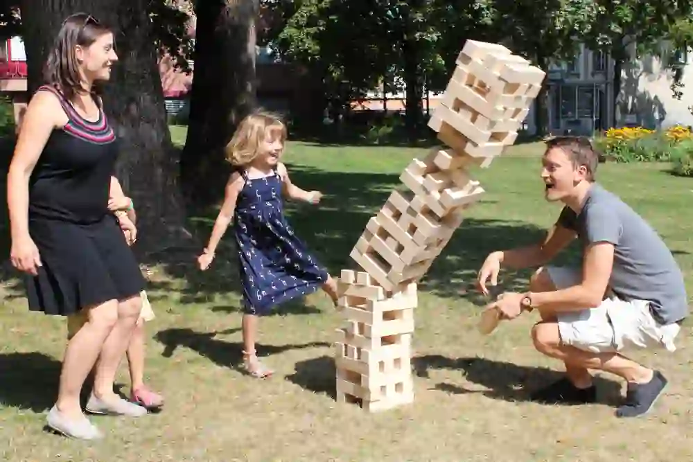 Eine Familie spielt lachend ein riesiges Jenga-Spiel im Freien; die kindliche Begeisterung und die gespannte Erwartung beim Kippen des Turms schaffen eine Atmosphäre ausgelassener Freude und sommerlicher Leichtigkeit.