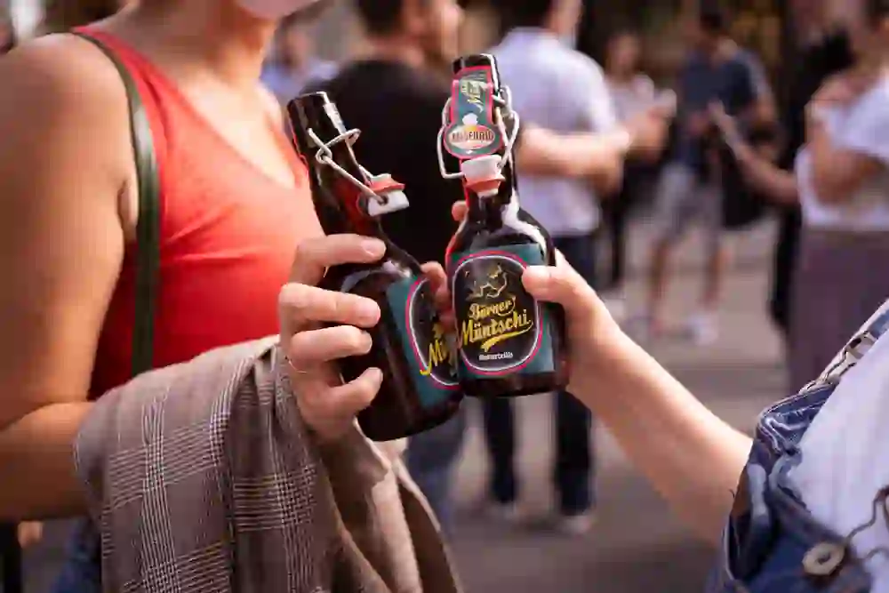 La photo montre deux mains qui tiennent deux bouteilles de bière différentes avec une fermeture à étrier. Les étiquettes sur les bouteilles indiquent qu'il s'agit de bières locales ou artisanales. En arrière-plan, on distingue une scène animée avec des personnes qui participent au Berner Biertour. La scène semble détendue et conviviale.