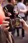La photo montre deux mains qui tiennent deux bouteilles de bière différentes avec une fermeture à étrier. Les étiquettes sur les bouteilles indiquent qu'il s'agit de bières locales ou artisanales. En arrière-plan, on distingue une scène animée avec des personnes qui participent au Berner Biertour. La scène semble détendue et conviviale.