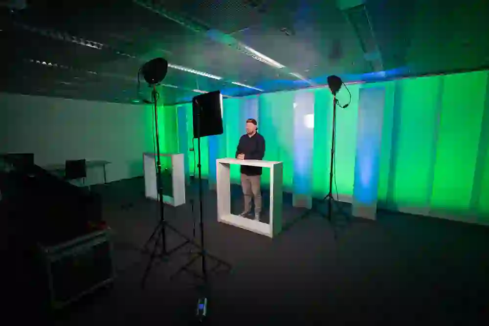 Un uomo concentrato è in piedi davanti a un leggio in uno studio moderno con illuminazione verde, circondato da luci professionali e apparecchiature di registrazione.