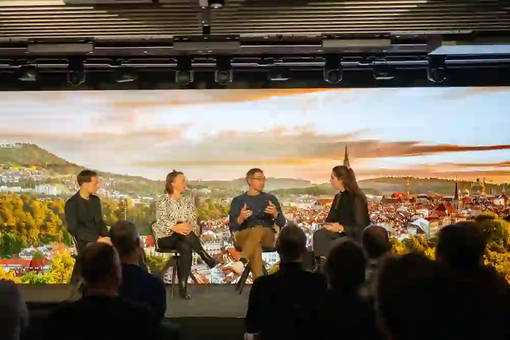 Auf dem Bild ist eine Diskussionsrunde vor einer Stadt-Kulisse bei Sonnenuntergang zu sehen. Vier Personen unterhalten sich engagiert, umgeben von einem interessierten Publikum, was eine inspirierende und dynamische Stimmung erzeugt.