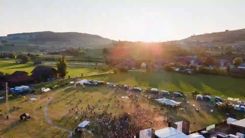 Das Bild zeigt das Stäcketöri Festival in Zäziwil aus der Luft, eingetaucht in goldenes Abendlicht. Die Sonne sinkt hinter sanften Hügeln, während Hunderte von Menschen ausgelassen feiern – ein magischer Sommerabend voller Musik, Freiheit und Gemeinschaft.