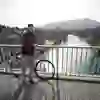 Ein lächelnder Mann steht an einem sonnigen Tag auf einer Brücke, die über einen klaren, türkisfarbenen Fluss führt. Mit seinem Fahrrad geniesst er die Aussicht auf die malerische Stadt Bern.
