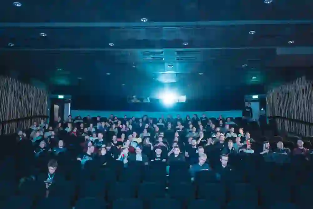 A full cinema auditorium with an eager audience, bathed in a soft blue light from the projector. The atmosphere is expectant and lively - you can feel the shared joy of film and the special atmosphere of a festival.