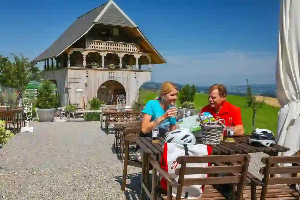 Auf diesem Foto geniesst ein Paar eine Pause auf der sonnigen Terrasse vom Hofladen Wielandleben in Röhtenbach, mit Blick auf eine malerische Landschaft. Sie sind in ein Gespräch vertieft, umgeben von rustikalen Holzmöbeln und Fahrradhelmen auf dem Tisch, was auf die E-Bike Tour im Emmental hindeutet. Die Kulisse mit einem charmanten Holzgebäude im Hintergrund strahlt Ruhe und ländliche Schönheit aus, und die entspannte Haltung des Paares vermittelt ein Gefühl von Zufriedenheit und Genuss im Moment.