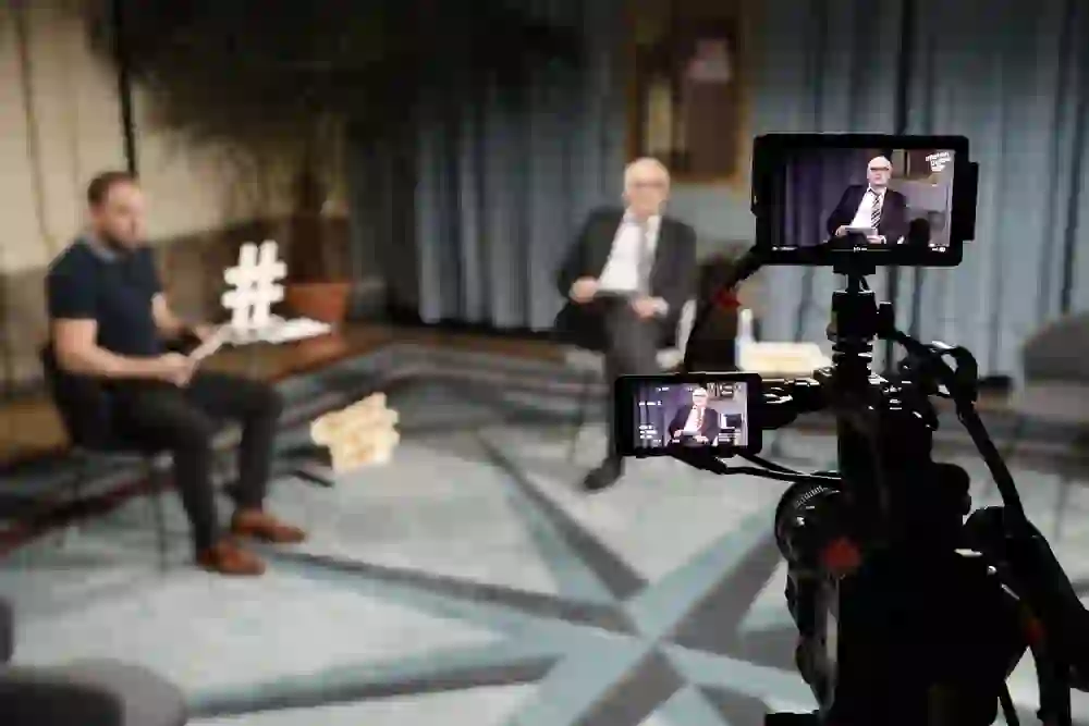 Das Bild zeigt eine Videoaufnahme in einem Innenraum: Eine Person ist verschwommen im Vordergrund sichtbar, während eine andere,  scharf fokussiert, im Hintergrund sitzt. Der Blick ist durch die Kamera gerichtet, die das Geschehen aufnimmt. Auf einem Display ist das Kamerabild zu sehen.