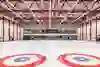 Une grande salle vide avec des cercles rouges et bleus distinctifs sur le sol, probablement pour le curling. L'atmosphère semble calme et prête pour la compétition.