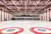 Una grande sala vuota con i caratteristici cerchi rossi e blu sul pavimento, presumibilmente per il curling. L'atmosfera è calma e pronta per la competizione.