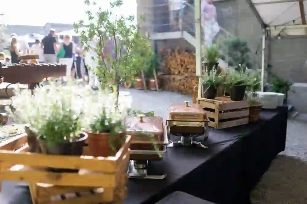 Elegante buffet all'aperto con erbe, piatti caldi e stoviglie. Ospiti che socializzano sullo sfondo. La scena è estiva, vivace e invitante, piena di divertimento e di incontri rilassati.