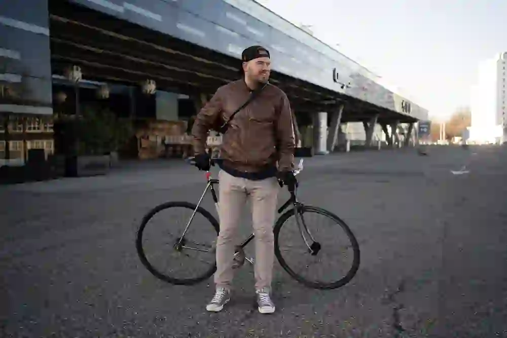 Brian Ruchti, vestito in modo casual, si trova in una piazza vuota in sella alla sua bicicletta, con un'architettura moderna sullo sfondo. Il suo sorriso rilassato e il suo atteggiamento disinvolto trasmettono un senso di libertà e spensieratezza.