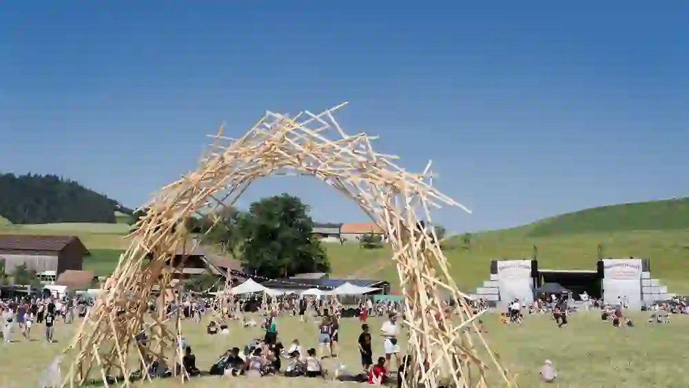 Das Bild zeigt das Stäcketöri Festival in Zäziwil bei strahlendem Sonnenschein. Unter einem kunstvollen Holzbogen sammeln sich Besucher:innen auf der Wiese, entspannen, lachen, geniessen – ein Fest der Freiheit, Kreativität und sommerlichen Leichtigkeit.