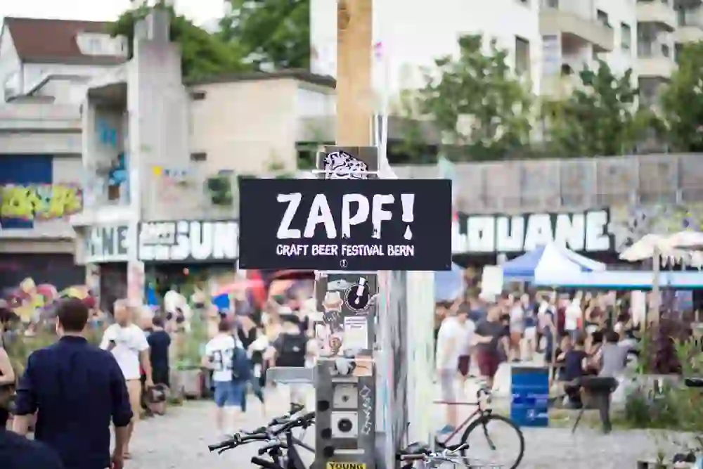 Das Bild fängt das lebhafte Treiben eines Craft Beer Festivals in Bern ein, mit einem Schild, das "ZAPF! Craft Beer Festival Bern" ankündigt, und einer entspannten Menschenmenge im Hintergrund, die das urbane, kulturelle Flair genießt.