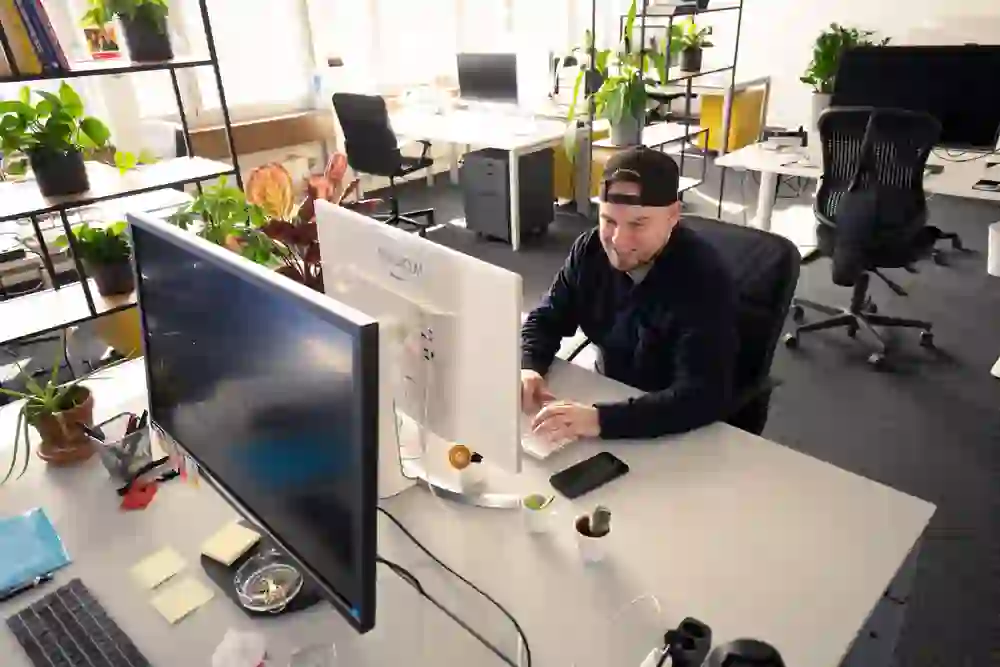 Brian Ruchti, con un berretto, sorride mentre lavora al computer in una postazione moderna e luminosa. L'ambiente è vivacemente decorato con piante che creano un'atmosfera rilassata e amichevole.