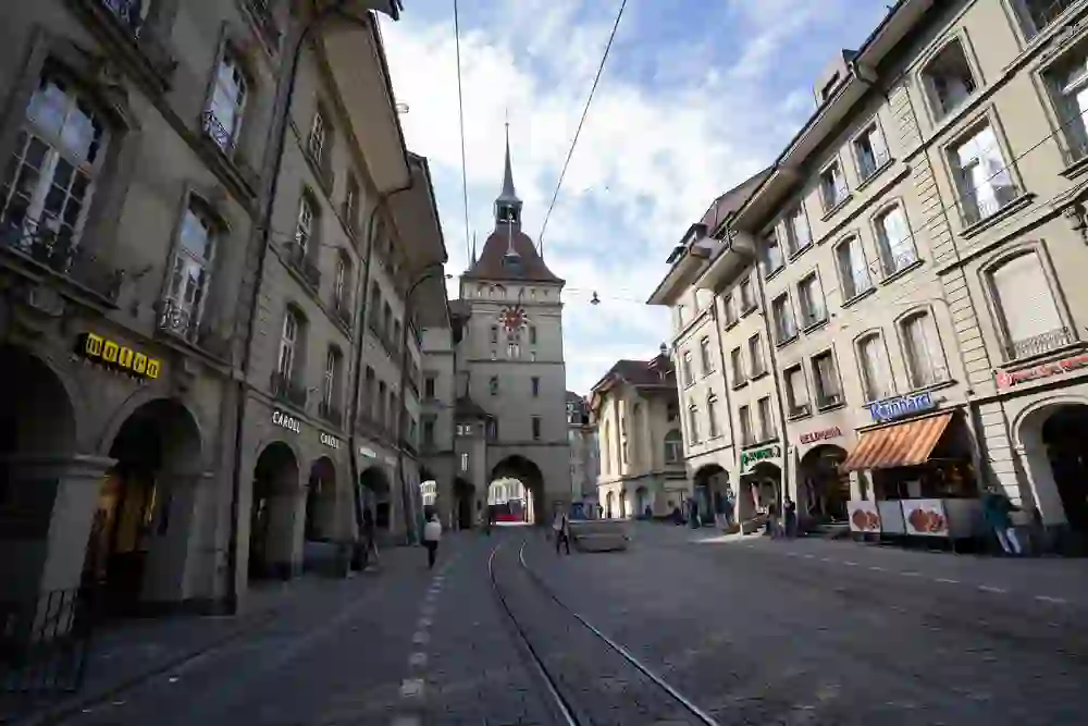 Das Bild zeigt eine lebhafte Straßenszene in einer europäischen Stadt mit historischer Architektur und einer Straßenbahnlinie. Menschen gehen ihrer Wege unter alten Gebäuden und Arkaden, was eine Atmosphäre von Alltagsleben und Geschichte schafft.