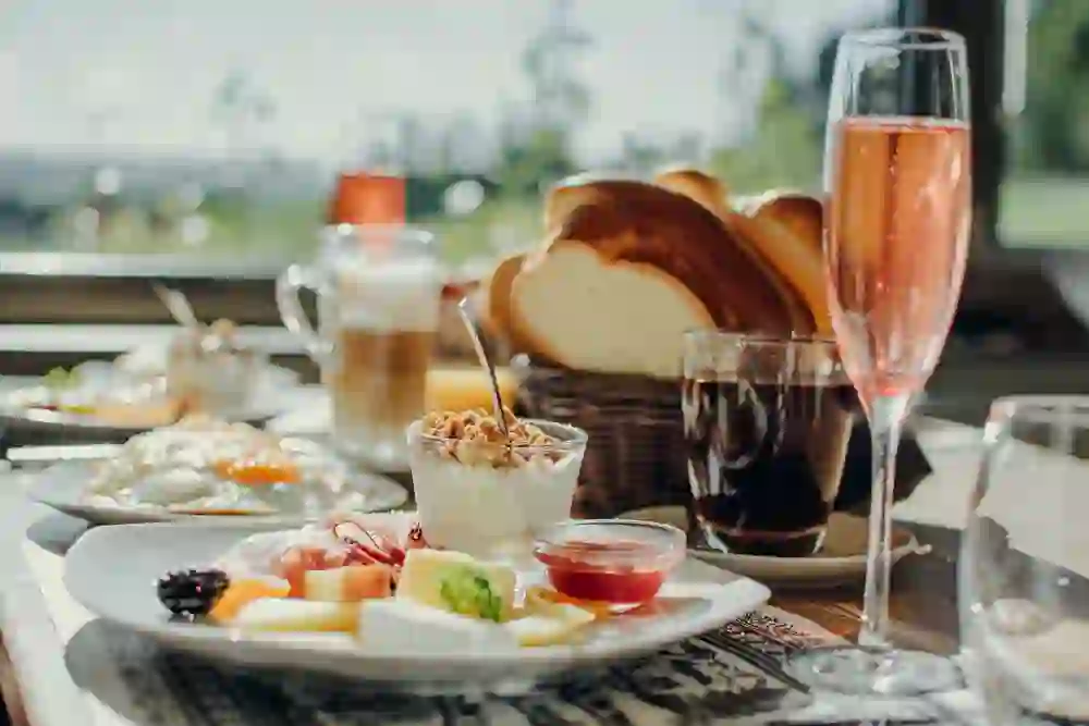 Das Bild zeigt ein reichhaltiges Frühstück, das in einer einladenden, sonnigen Atmosphäre serviert wird. Im Mittelpunkt steht ein Teller mit einer Auswahl an Käse, Aufschnitt, frischen Früchten und Marmelade. Daneben befindet sich ein Glas mit Joghurt und knusprigem Müsli. Im Hintergrund sind ein Latte Macchiato, ein Glas Cola, ein Glas Rosé-Sekt sowie ein Korb mit frischem Zopf-Brot zu sehen.  Die warme natürliche Beleuchtung verleiht dem Bild eine besonders gemütliche und genussvolle Stimmung. Es könnte sich um ein Frühstück in einem traditionellen Schweizer Restaurant oder einer Schaukäserei im Emmental handeln, das regionale Spezialitäten präsentiert.