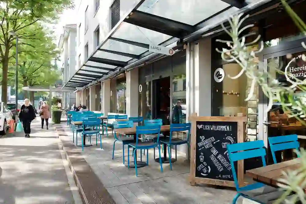 Die Aufnahme zeigt die gemütliche Außenterrasse eines Cafés oder Restaurants vor einem Hotel. Türkisfarbene Stühle und Holztische laden Passanten unter einem modernen Glasdach zum Verweilen ein. Ein handbeschriftetes Willkommensschild vermittelt eine einladende, freundliche Atmosphäre. 🌿☕