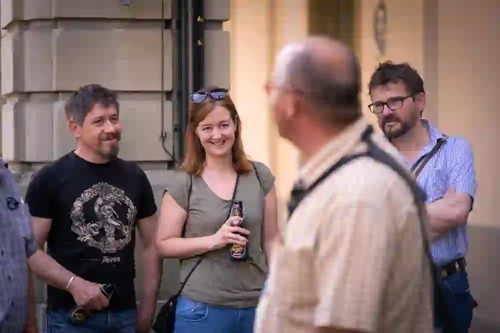 Sur cette photo, un petit groupe d'adultes regarde avec attention et amusement un guide de la ville qui se tient en dehors de la photo. Ils semblent détendus et intéressés ; une femme sourit en tenant une bière et les hommes à côté d'elle écoutent avec le même intérêt. L'ambiance est détendue et amicale.