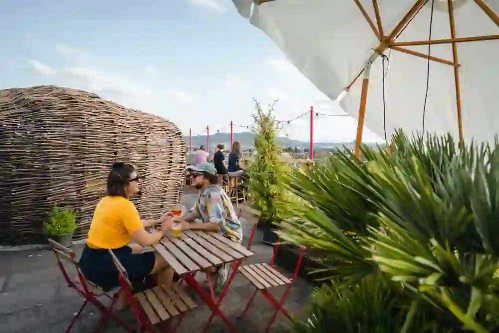 Auf dem Foto ist die entspannte Dachterrasse von der Bar 8 in Bern zu sehen, die zu gemütlichen Gesprächen bei einem Drink einlädt. Die Szene ist geprägt von einer lässigen, freundlichen Atmosphäre. Ein Paar geniesst die gemeinsame Zeit an einem kleinen Tisch, umgeben von Pflanzen und unter dem Schutz eines grossen Sonnenschirms. Im Hintergrund sorgt die verwobene Weidenstruktur für eine künstlerische Abgrenzung, während andere Gäste die Aussicht und das schöne Wetter geniessen.