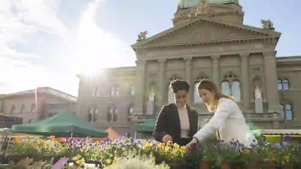 Zwei Frauen betrachten fröhlich blühende Blumen an einem Marktstand auf dem Bundesplatz in Bern. Im Hintergrund steht das Bundeshaus im warmen Morgenlicht, umgeben von bunten Marktzelten. Die Szene vermittelt urbanes Lebensgefühl, Nahbarkeit und die genussvolle Seite der Stadt.