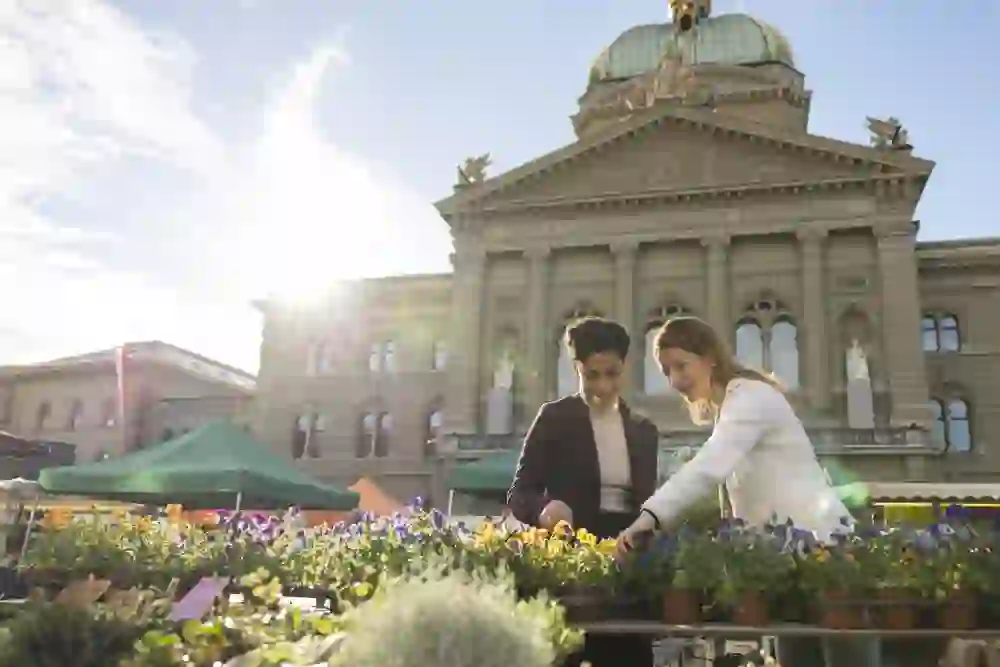 Zwei Frauen betrachten fröhlich blühende Blumen an einem Marktstand auf dem Bundesplatz in Bern. Im Hintergrund steht das Bundeshaus im warmen Morgenlicht, umgeben von bunten Marktzelten. Die Szene vermittelt urbanes Lebensgefühl, Nahbarkeit und die genussvolle Seite der Stadt.