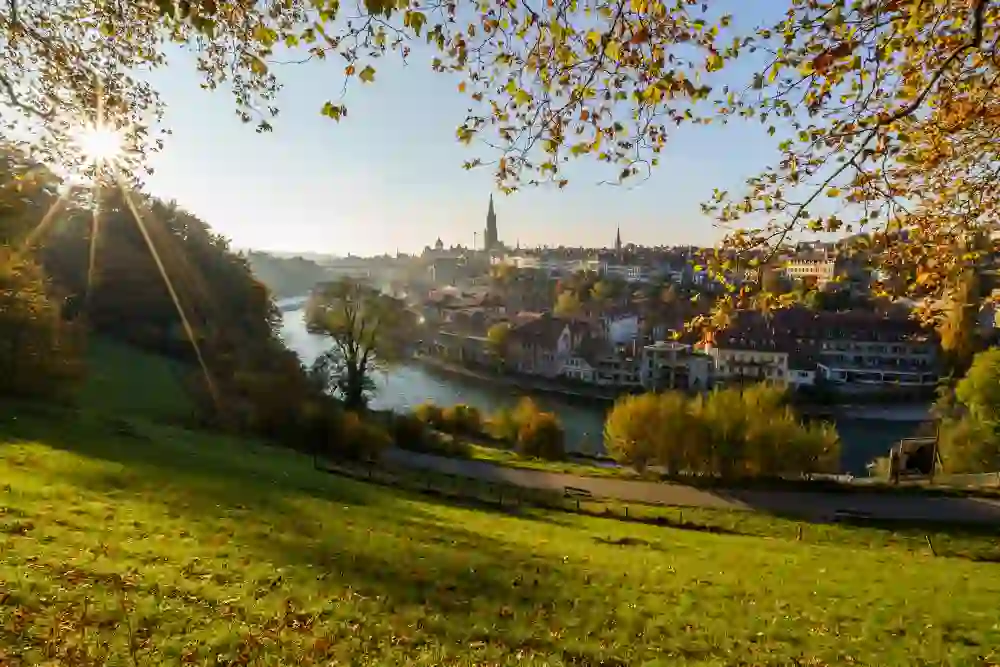 A view across Bern’s old town, with the River Aare, the silhouette of the cathedral and autumn-coloured trees in the foreground. Sunbeams filter through the foliage, bathing the landscape in warm light. The atmosphere is peaceful, inviting and autumnal.