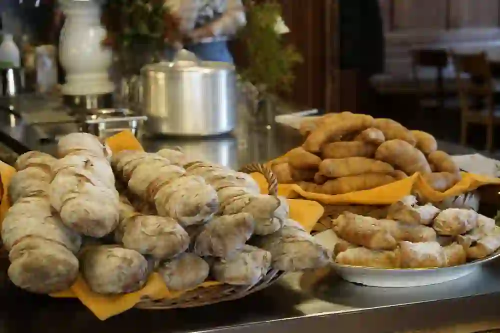 Das Bild zeigt eine Auswahl an Backwaren auf einer Theke, darunter Ciabatta-Brote, Croissants und gefüllte Teigtaschen, ansprechend auf Körben mit orangefarbenen Tüchern drapiert. Im Hintergrund ist ein Teil der Café-Einrichtung und eine Person, die im Service arbeitet, verschwommen sichtbar.