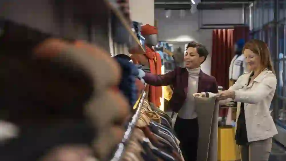 Zwei Frauen stöbern gemeinsam durch ein hochwertig sortiertes Modegeschäft in der Berner Altstadt. Sie wirken entspannt und lachen, während sie Kleidungsstücke begutachten – umgeben von farbenfrohen Stoffen, stilvoller Einrichtung und urbaner Atmosphäre.