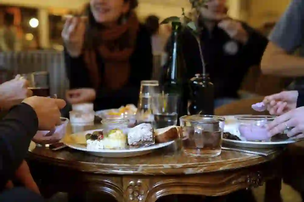 Das Foto zeigt eine gesellige Runde von Personen, die an einem Tisch sitzen und gemeinsam essen. Auf dem Tisch befinden sich verschiedene Gerichte und Getränke, darunter Käse, Brot, eine weiße Kaffeetasse und Gläser, wahrscheinlich mit Wasser oder Tee. Im Hintergrund ist eine Frau zu sehen, die lächelt und anscheinend eine Unterhaltung führt. Die Szene spielt sich in einem warm beleuchteten Raum ab, der eine heimelige Atmosphäre ausstrahlt.