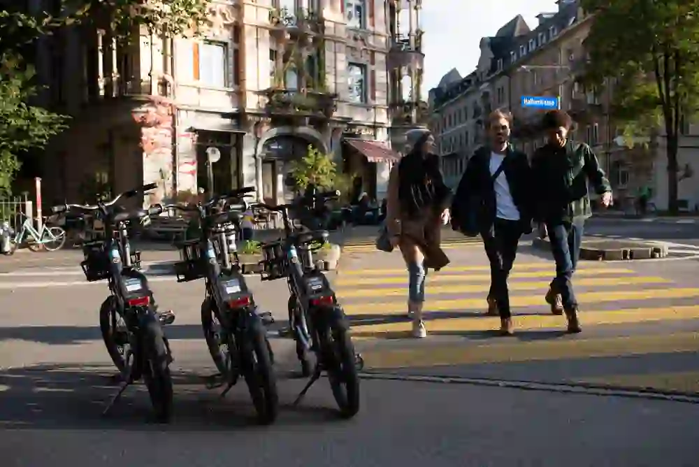 Three friends smile as they cross a busy city street past a row of parked bikes, captured in a moment of lightness and connection, accompanied by the urban charm and freedom offered by a PubliBike day pass.