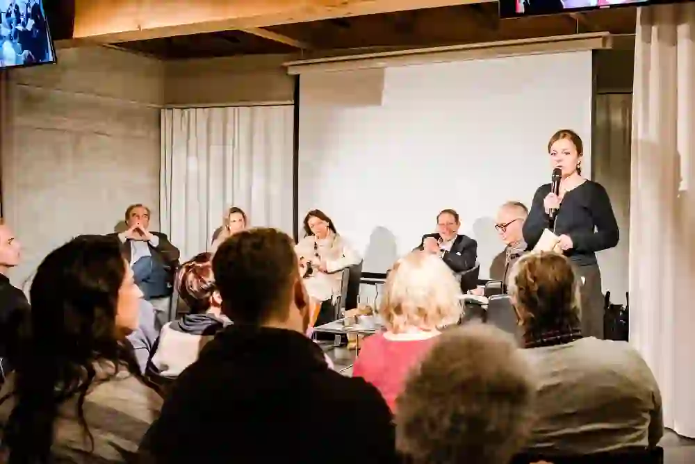 Das Bild zeigt eine Podiumsdiskussion in einem belebten Veranstaltungsraum. Eine Sprecherin steht vor dem Publikum, das aufmerksam zuhört. Die Stimmung ist ernst und engagiert, reflektiert durch das interessierte Publikum und die konzentrierten Panel-Teilnehmer.