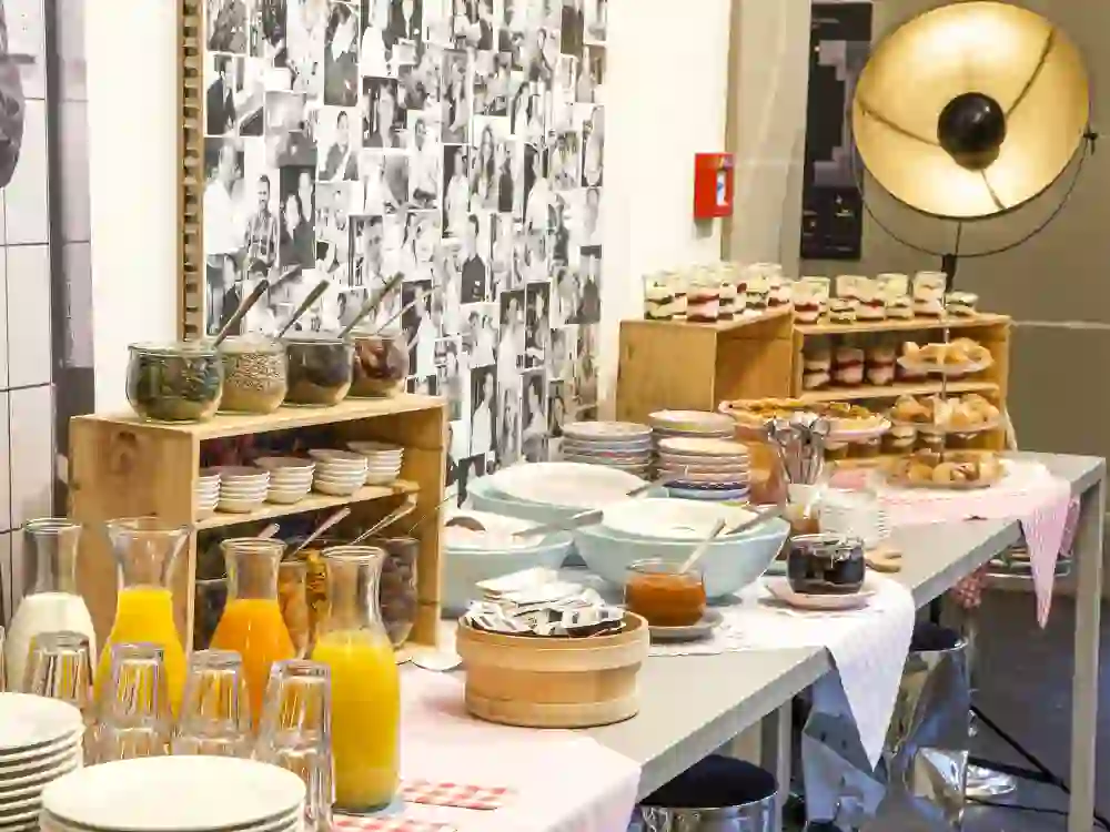 Das Bild zeigt ein einladendes Brunch-Buffet im Restaurant "Toi et Moi". Mit einer lebhaften Wand voller Porträts als Hintergrund, strahlt das Arrangement von frischen Backwaren, Gläsern mit farbenfrohen Säften und Schalen mit Müsli eine heimelige und gastfreundliche Atmosphäre aus, die zum Verweilen und Genießen einlädt.