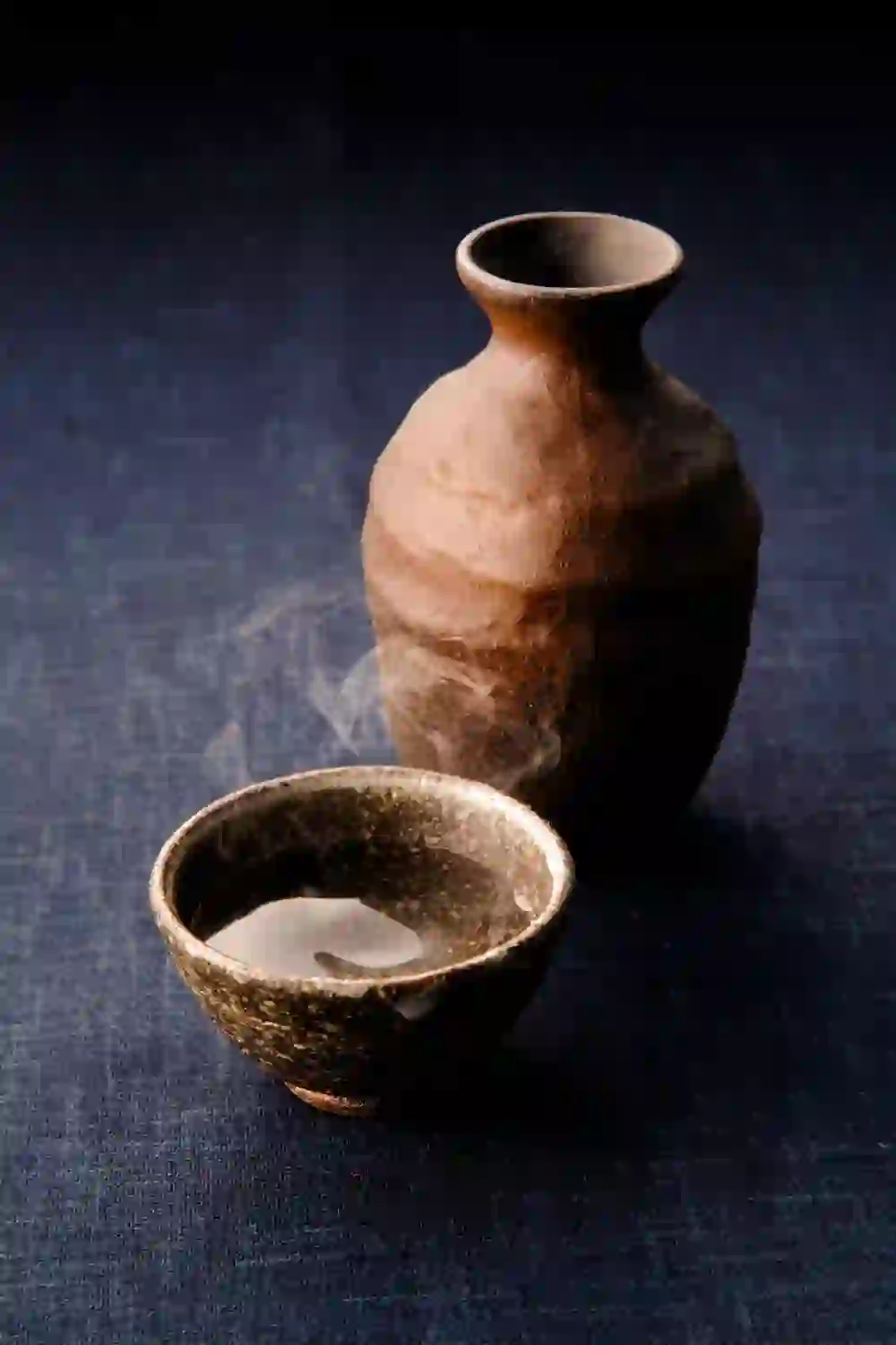 L'image montre une bouteille de saké traditionnelle et un bol de saké fumant. La céramique rustique et la vapeur qui s'élève créent une atmosphère chaleureuse et méditative qui invite à savourer et à se détendre. La scène respire le calme et la simplicité.