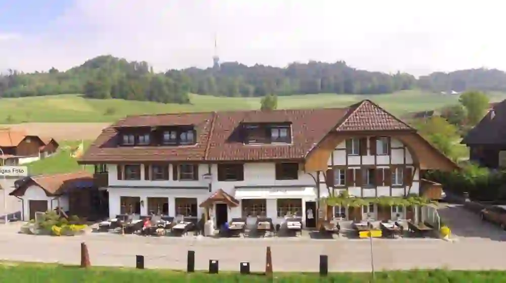 The picture shows the "Gasthof Alpenblick" in Ferenberg, a traditional Swiss inn with an outdoor terrace. Tables are set up in front of the inn for guests to enjoy the view of the surrounding green landscape. The scenery is rural and peaceful, with a clear blue sky, creating a calm and relaxing atmosphere.