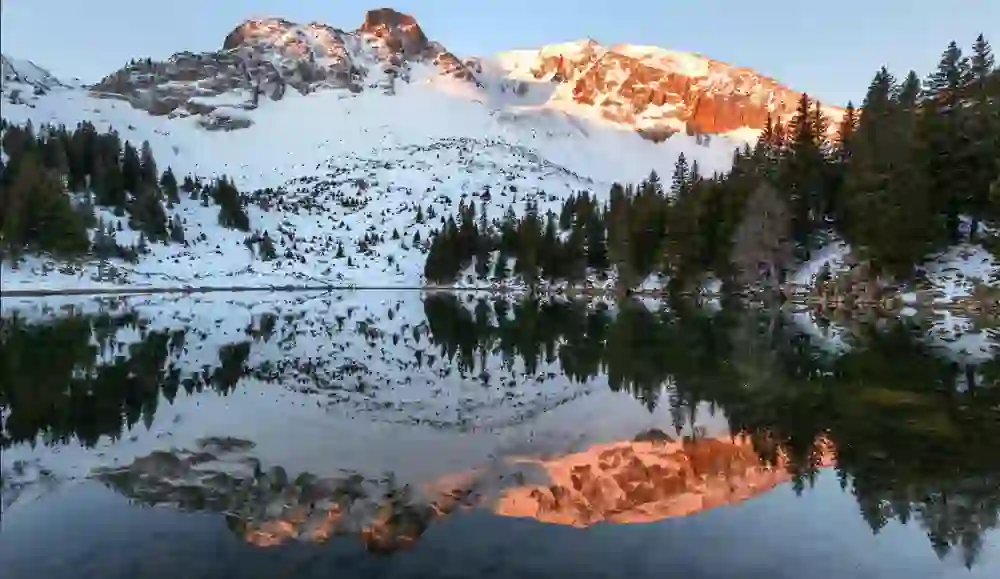 Das Bild zeigt einen ruhigen Bergsee, der die schneebedeckten Gipfel im sanften Licht des Sonnenuntergangs widerspiegelt. Die rötlich leuchtenden Berge und der klare Himmel vermitteln eine friedliche, majestätische Stimmung, die zum Innehalten einlädt.
