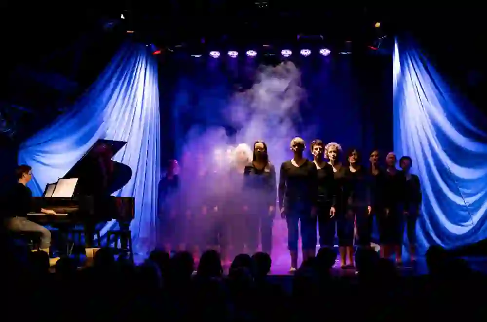 Das Bild zeigt einen Chor und eine Pianistin auf der Bühne des Kulturhof Schloss Köniz. Die dramatische Beleuchtung und der Nebel kreieren eine zauberhafte Stimmung. Die ernsten Gesichter der Sängerinnen deuten auf die Intensität des Moments hin, während das Publikum gebannt zuhört.