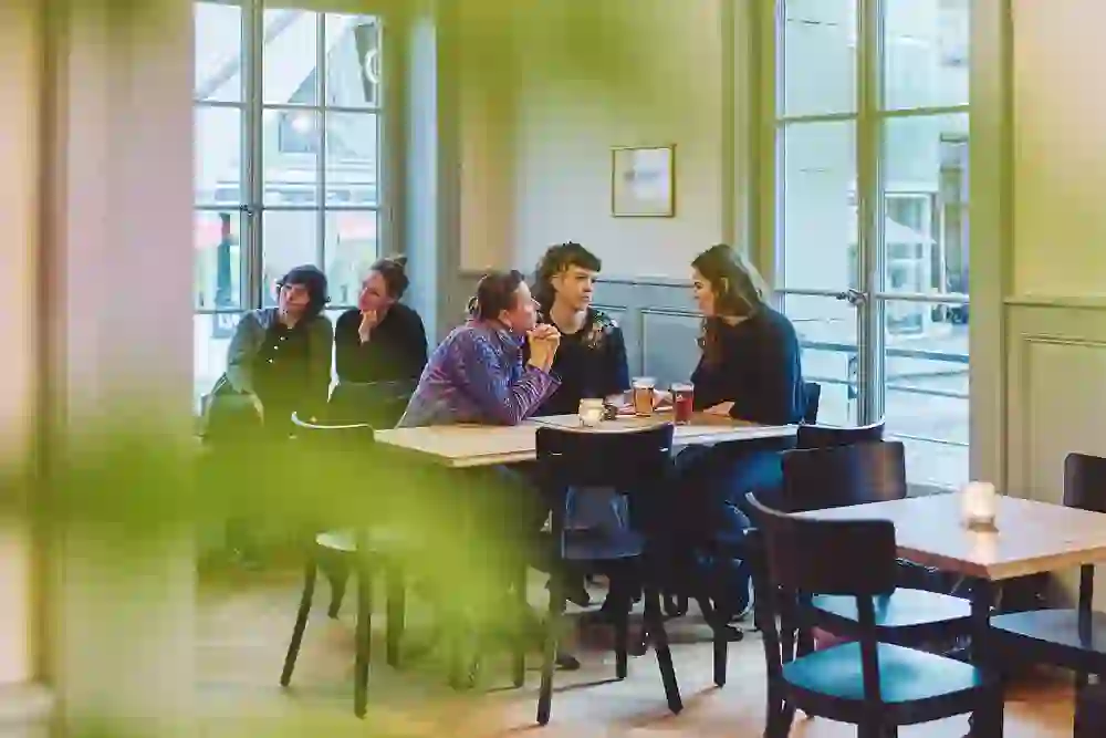 Das Bild zeigt eine gesellige Szene in einem stilvollen Café. Zwei Gruppen von Freunden genießen in entspannter Atmosphäre ihre Getränke, wobei die großen Fenster für natürliches Licht sorgen und die heimelige Stimmung unterstreichen.