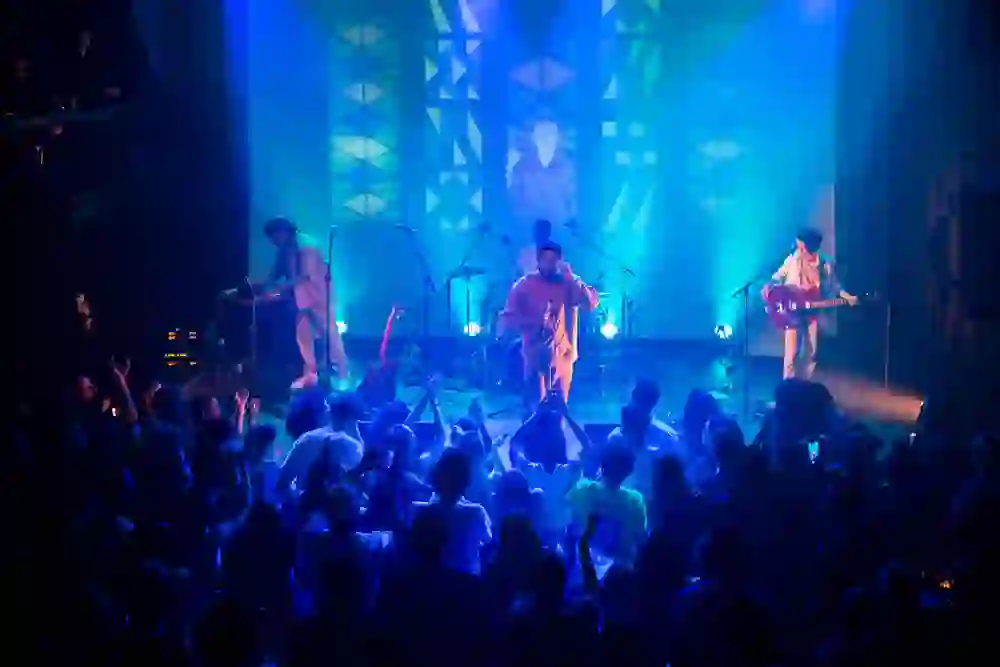 The picture shows an energetic concert scene in which the stage is bathed in blue light. The musicians perform in front of an enthusiastic audience, who cheer along with their hands raised. The powerful atmosphere and the connection between the artists and the audience convey the feeling of an intense, thrilling live experience.