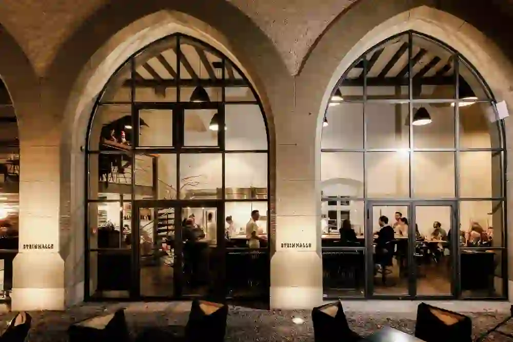 Das Bild zeigt die Außenansicht eines eleganten Restaurants mit großen, bogenförmigen Fenstern. Drinnen genießen Gäste in entspannter Atmosphäre ihr Essen. Die warme Beleuchtung strahlt Gemütlichkeit aus und lädt zum Verweilen und Genießen ein.